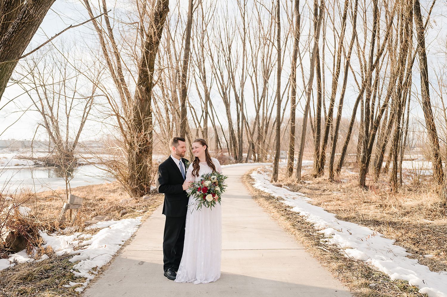 A winter wedding portrait on a path with bare trees and snowy ground.