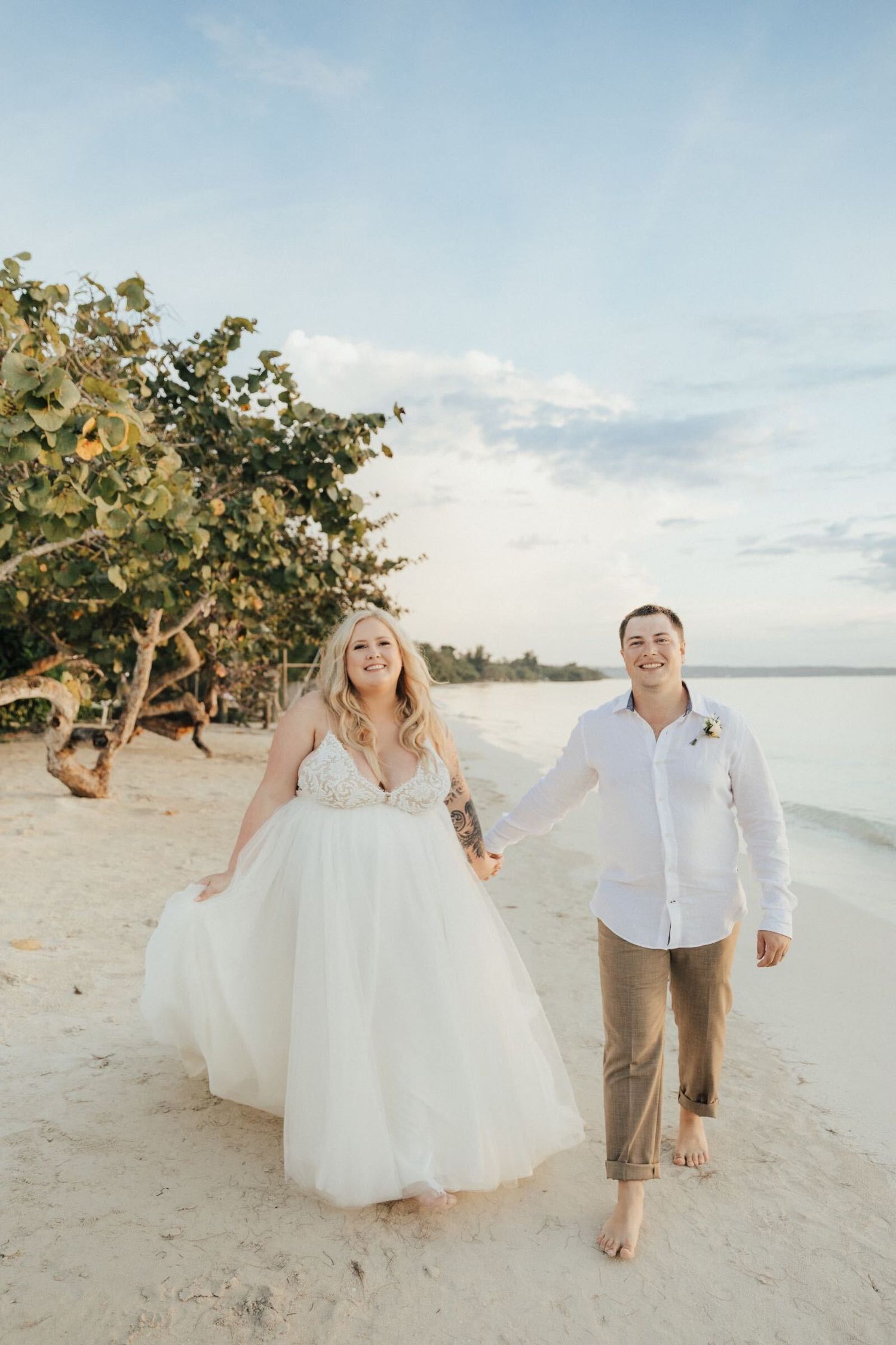 A couple in wedding attire walking along a tropical beach at sunset.