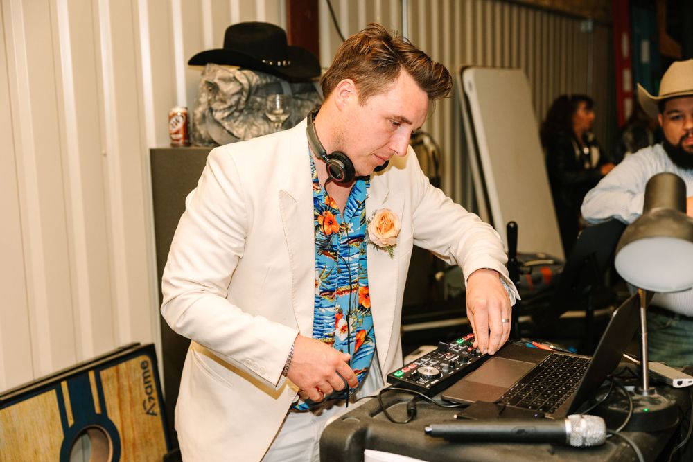 DJ wearing white blazer and floral shirt performing at event mixing console.