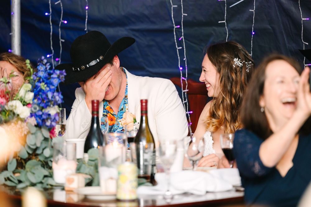 Wedding guests enjoy a lively reception dinner with blue floral decor and wine bottles on the table.