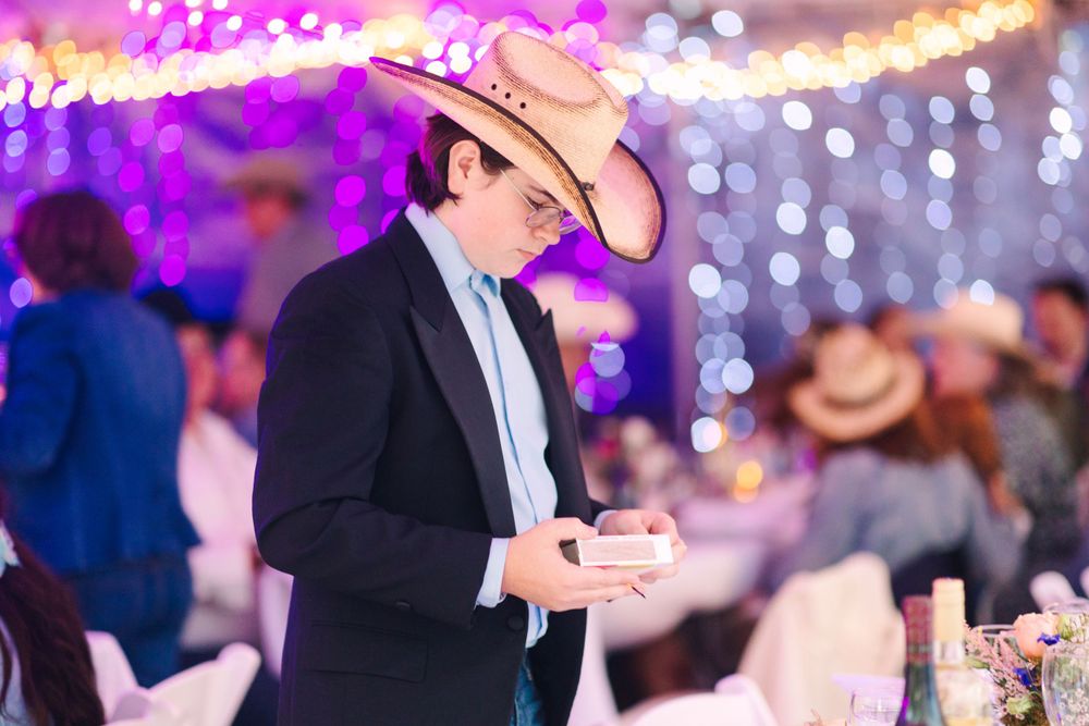A person in formal attire and cowboy hat checks their phone under purple party lighting.