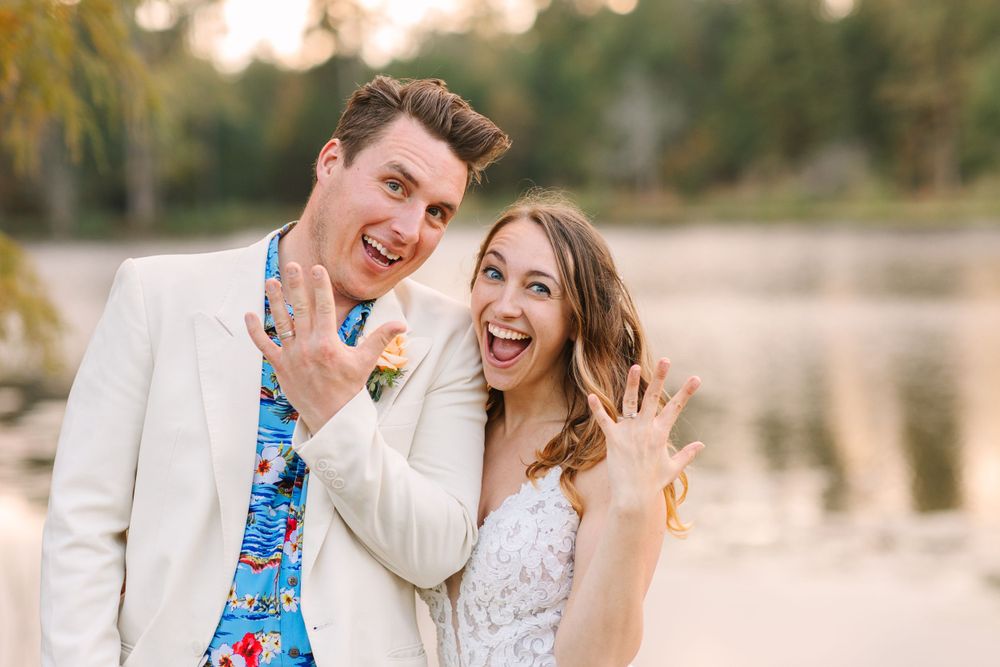 A joyful couple showing off their rings outdoors by a lake during a sunset engagement or wedding photo session.