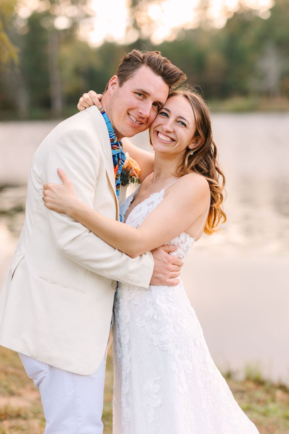 Happy couple embraces while wearing white wedding attire against sunset lake backdrop.