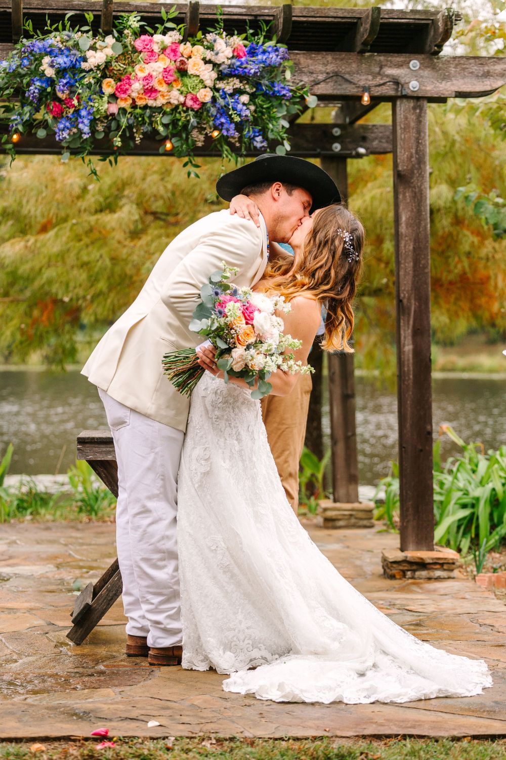 Couple shares romantic kiss under floral arch by scenic lake during autumn wedding.