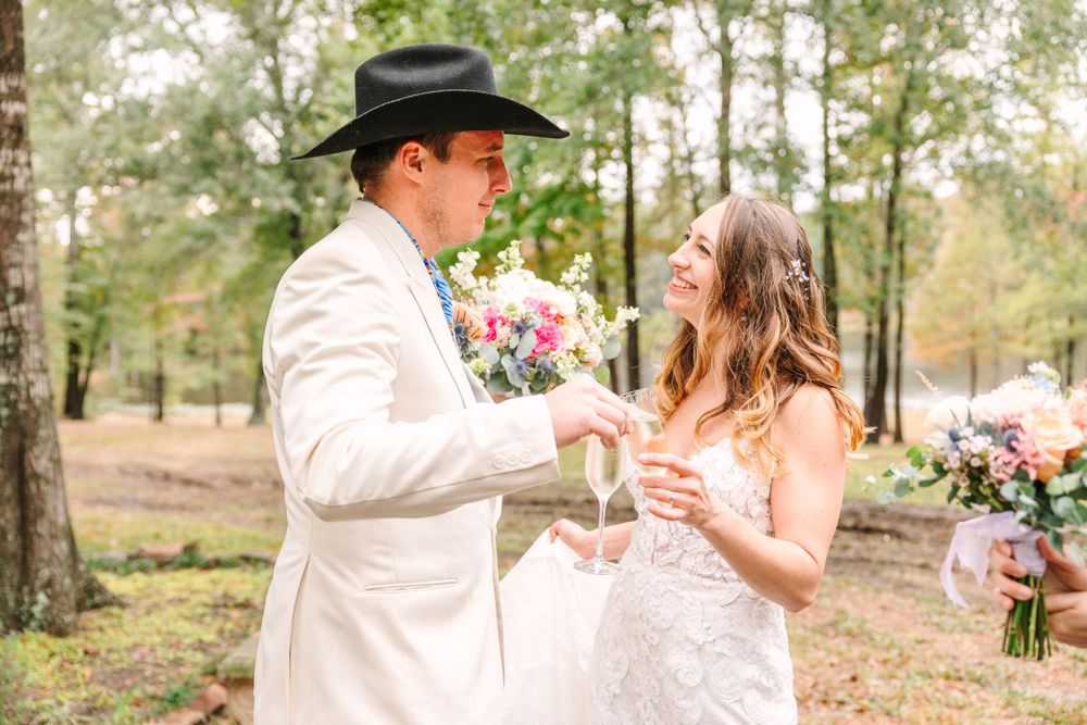 A couple in wedding attire sharing a toast outdoors, with the groom wearing a white suit and cowboy hat.