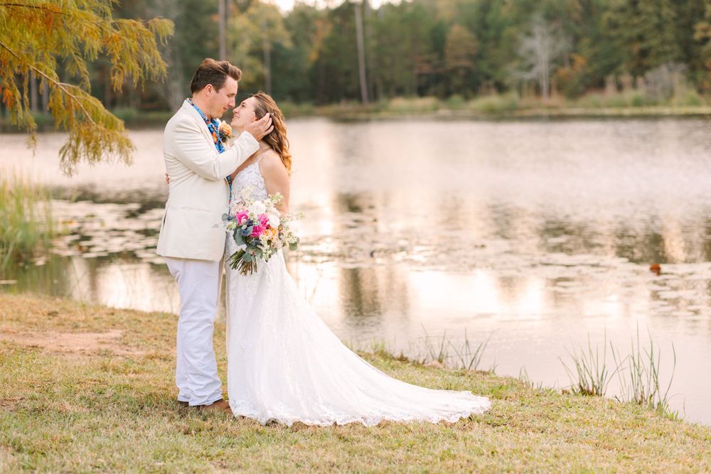Wedding couple embraces by tranquil lake during golden hour with trees in background.