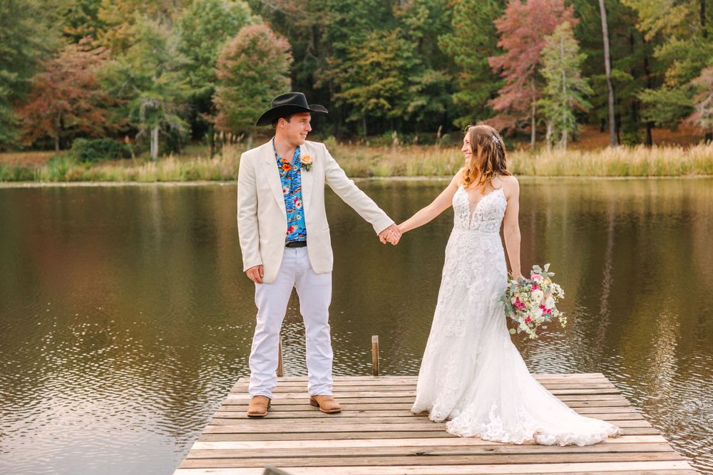 Newlyweds hold hands while standing on wooden dock with lake and fall foliage in background.
