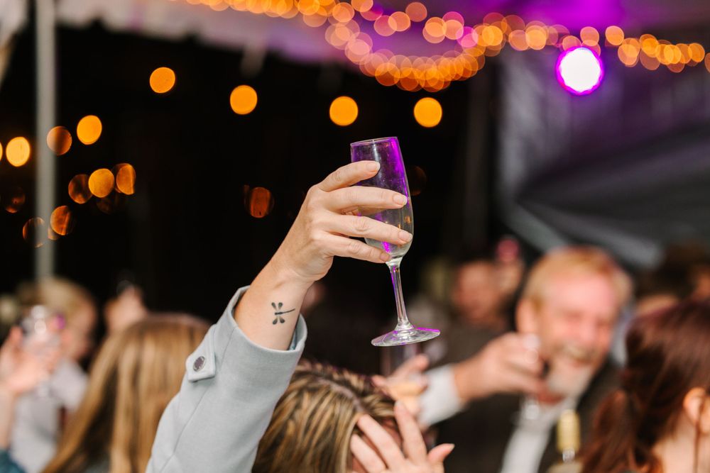 A hand raises a purple-tinted champagne glass during an evening celebration with bokeh lights.