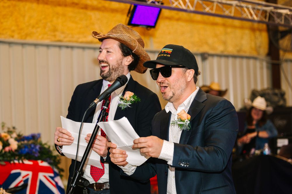 Two men in suits and cowboy attire energetically read from papers during a wedding celebration.