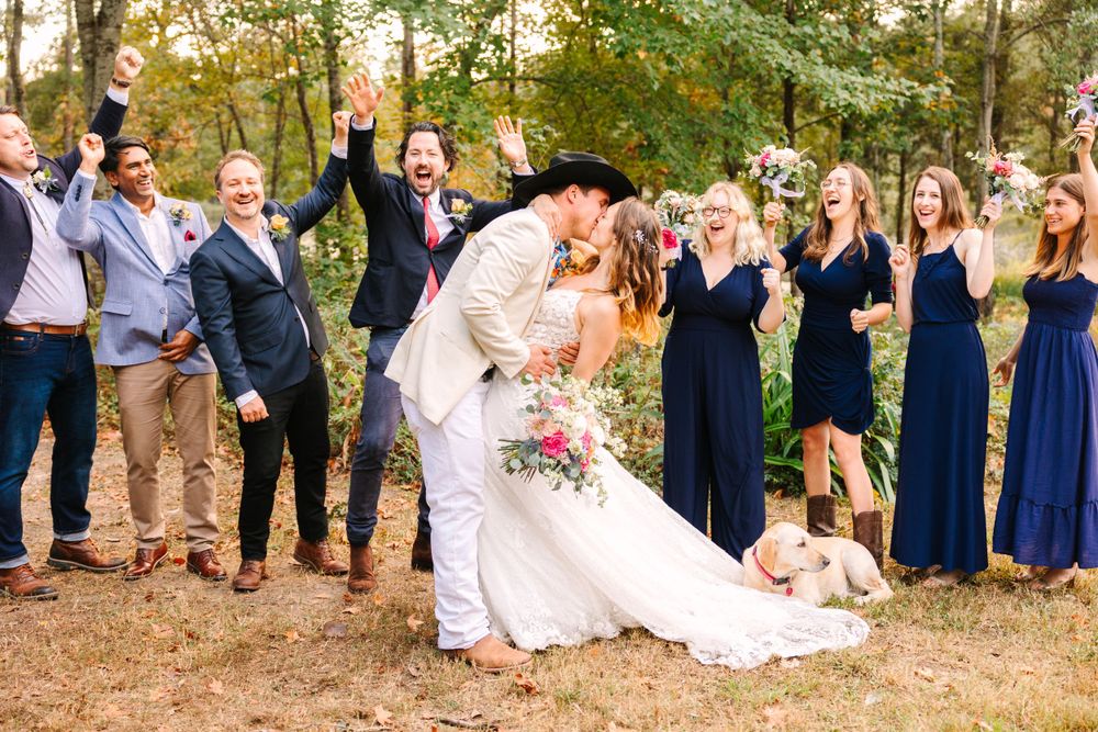 Wedding party celebrates with raised hands while couple shares romantic moment in center.