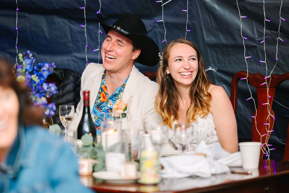 A couple at their reception table laughing together under decorative string lights.