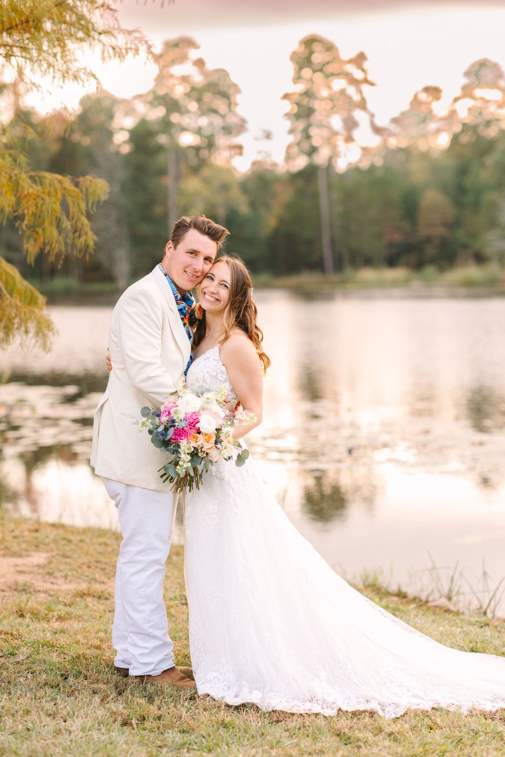 Couple poses by lakeside during autumn sunset with colorful bouquet and fall foliage.