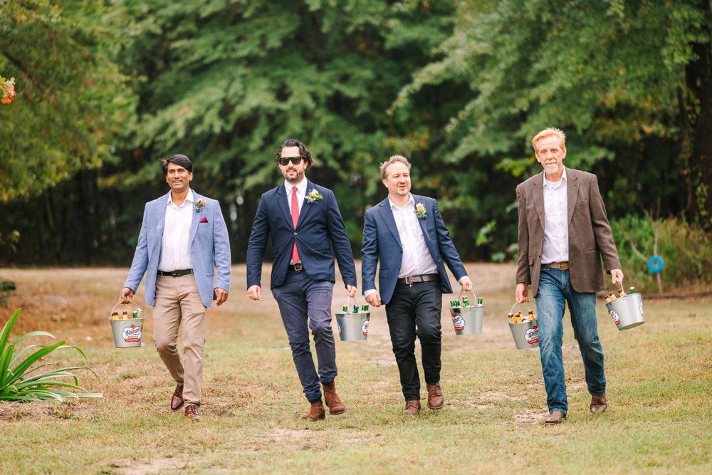 Four groomsmen walk together across a lawn carrying drinks in a casual outdoor wedding setting.