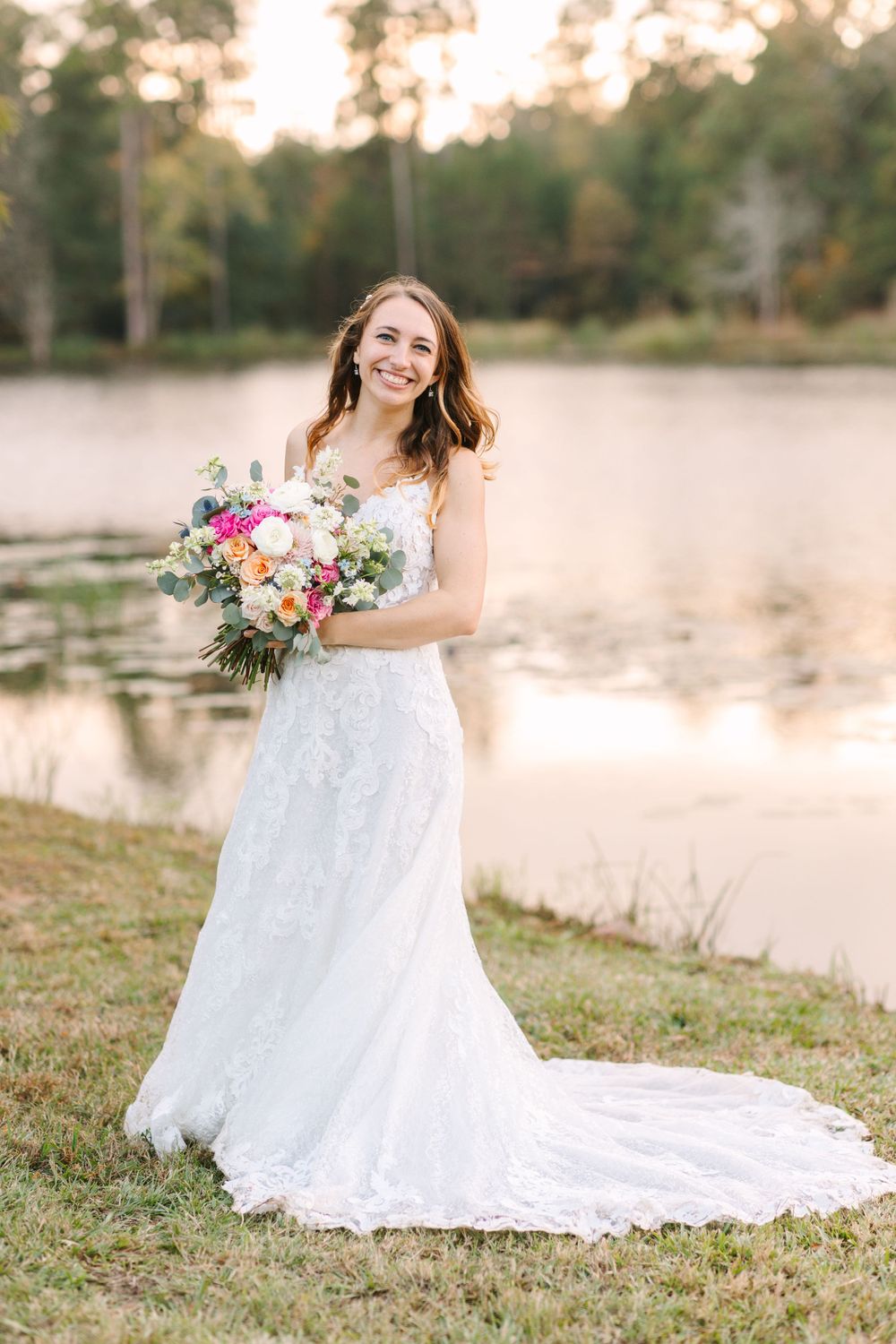 A bride in a white lace gown holds a colorful bouquet while posing near a lake at golden hour.