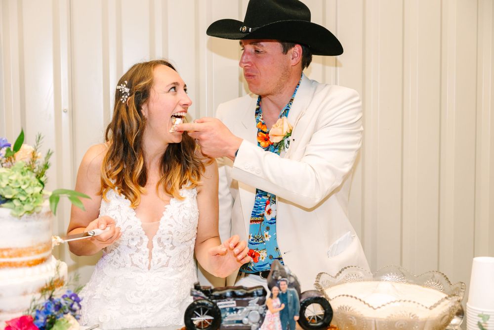 Couple shares cake at their wedding reception with a tiered floral cake in the background.