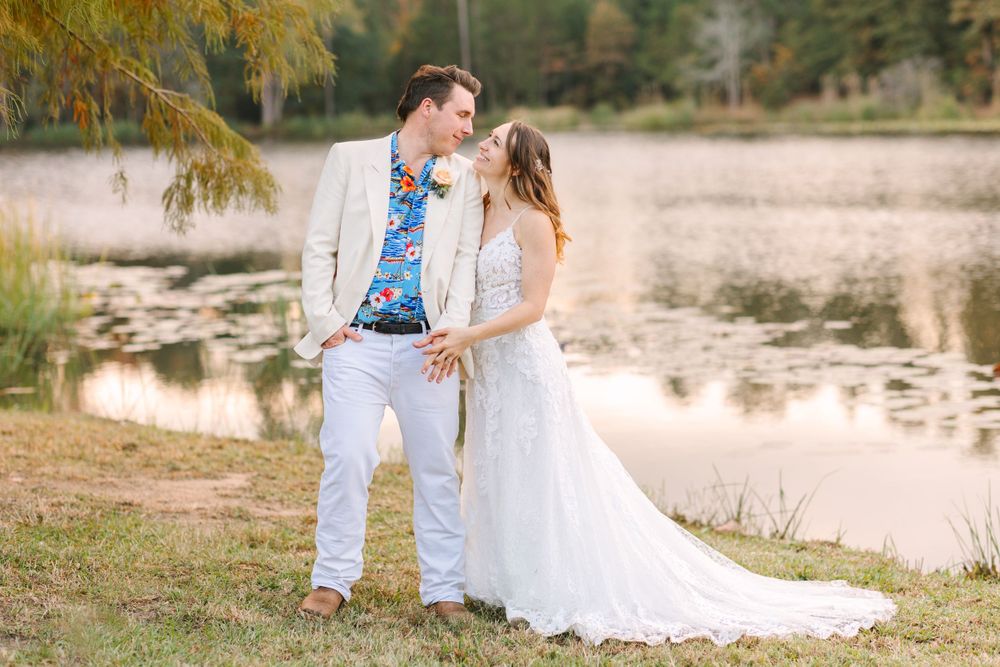 Couple in white wedding attire shares tender moment by lake with autumn trees in background.