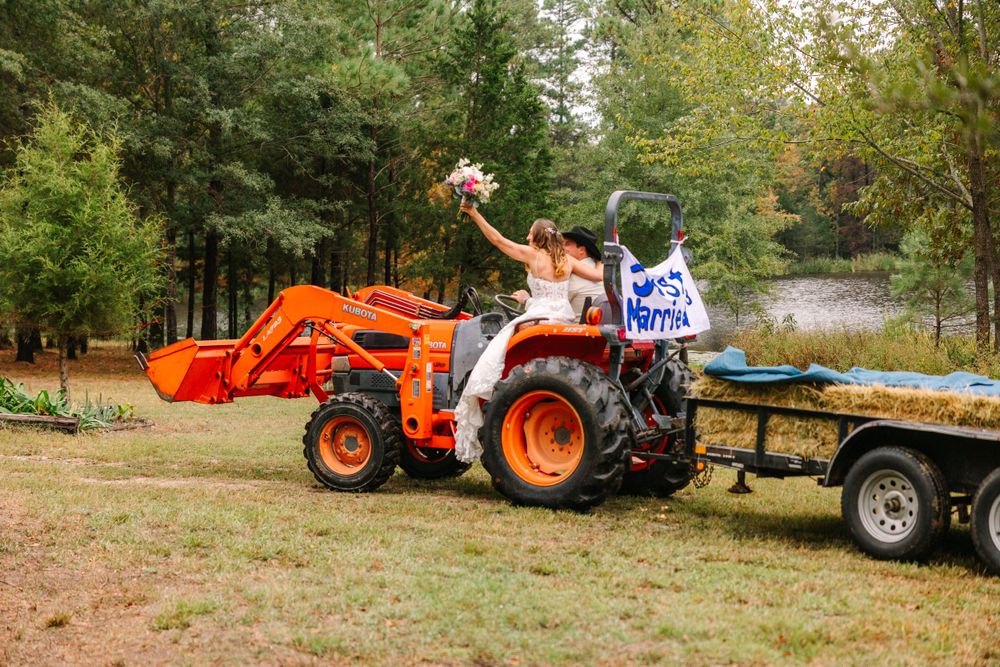 Couple celebrates on orange tractor with decorative signs at outdoor wedding venue.
