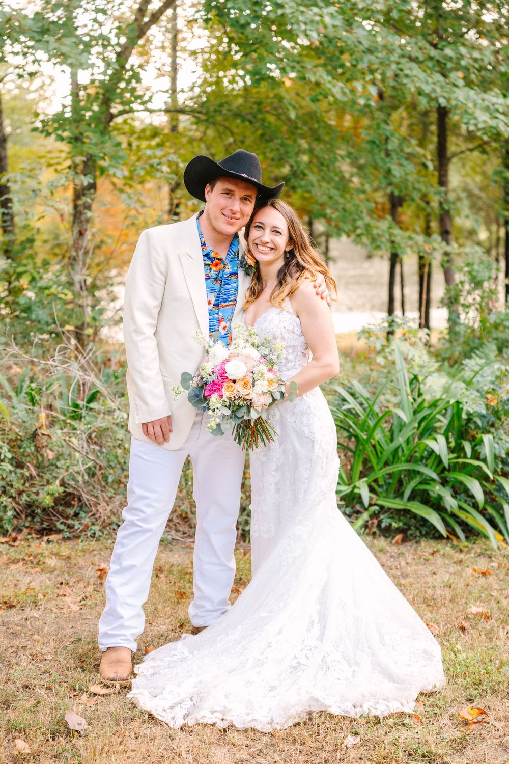 A couple poses outdoors in wedding attire with the groom in a white suit and black cowboy hat among autumn trees.