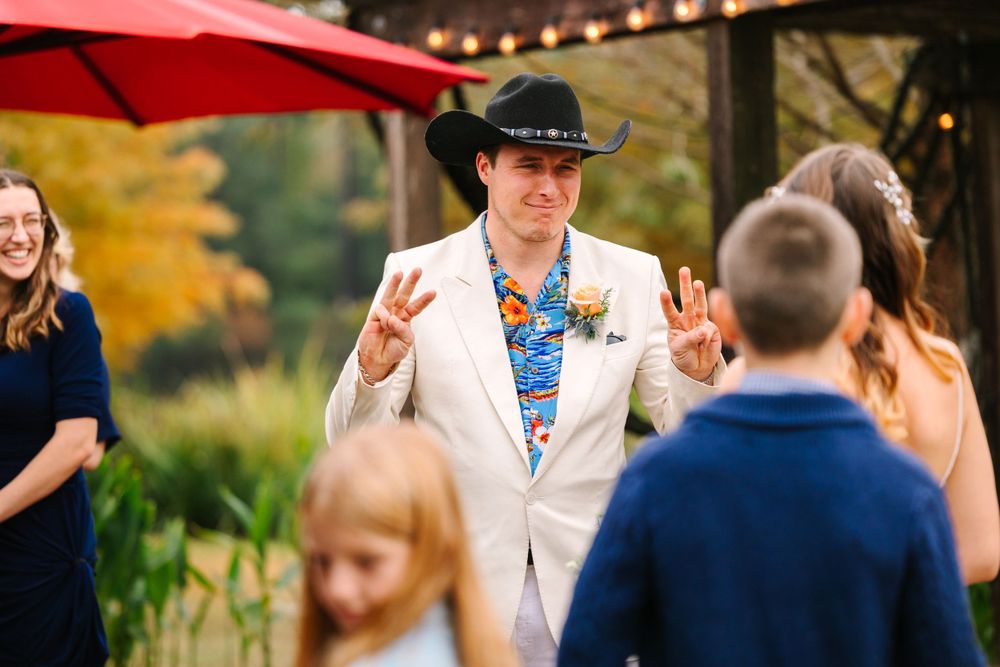 Person in white suit jacket and cowboy hat gestures during outdoor wedding ceremony under autumn trees.