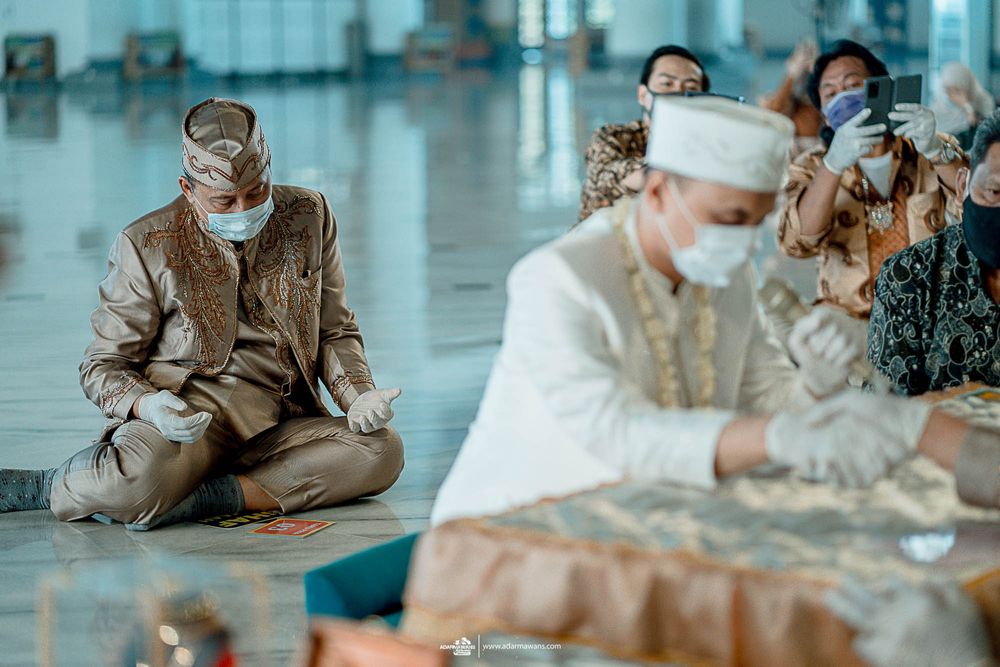 People wearing masks sit on the floor in a modern building during a ceremonial event.