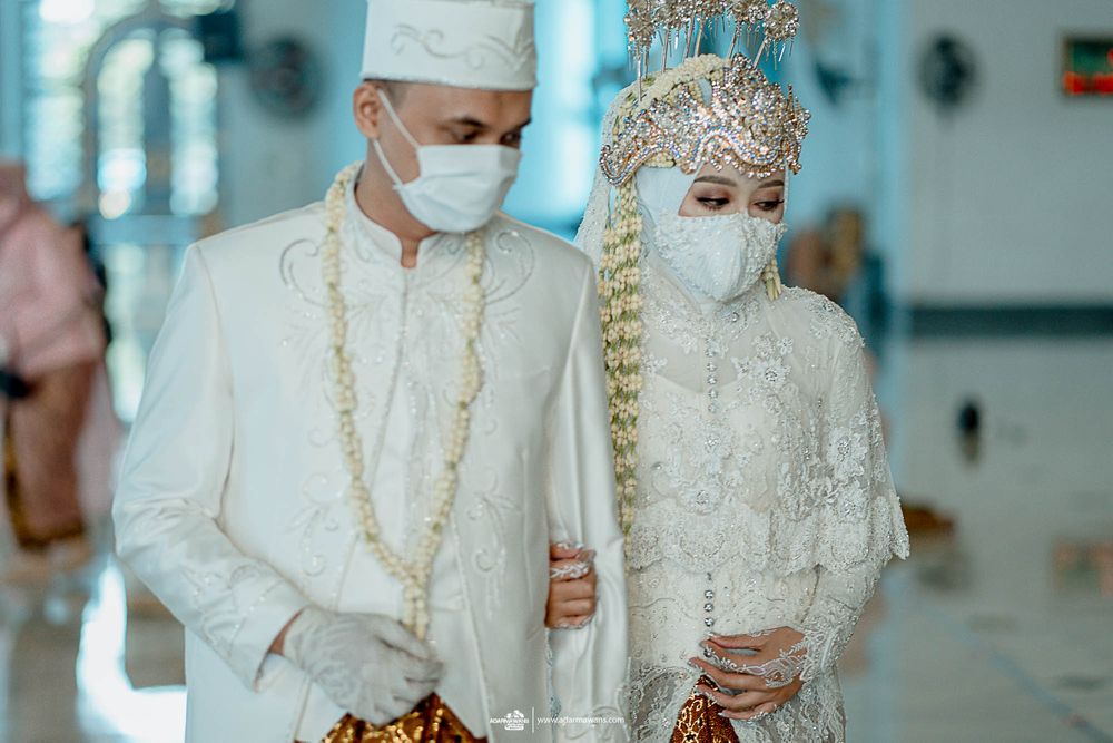 A couple in ornate white traditional wedding attire and face masks stand together.