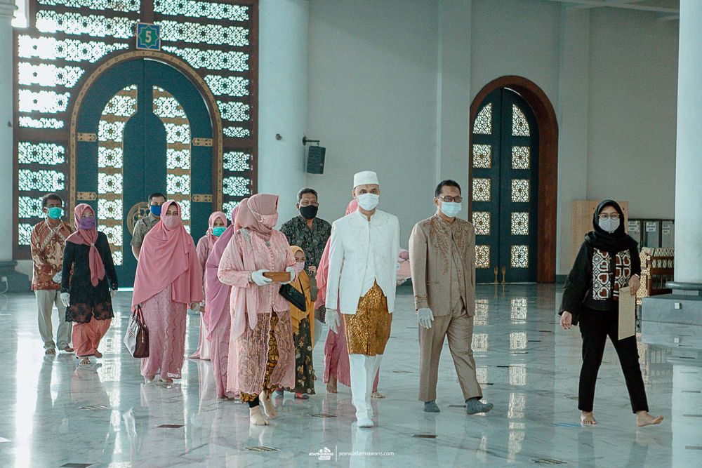 Group of worshippers wearing protective masks and traditional attire gathering inside a mosque's bright interior space.