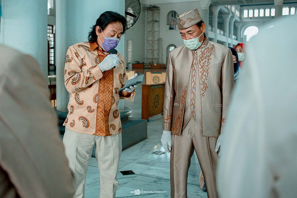 Two individuals in formal attire wearing face masks while discussing in a mosque interior.