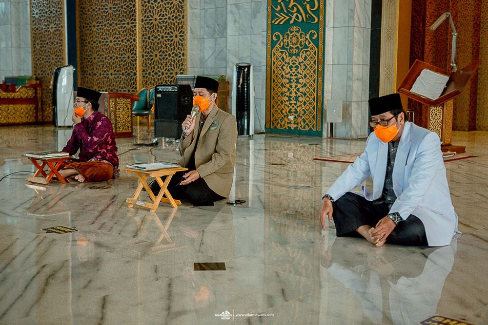 Three men sitting distanced on prayer mats inside a decorated mosque interior.
