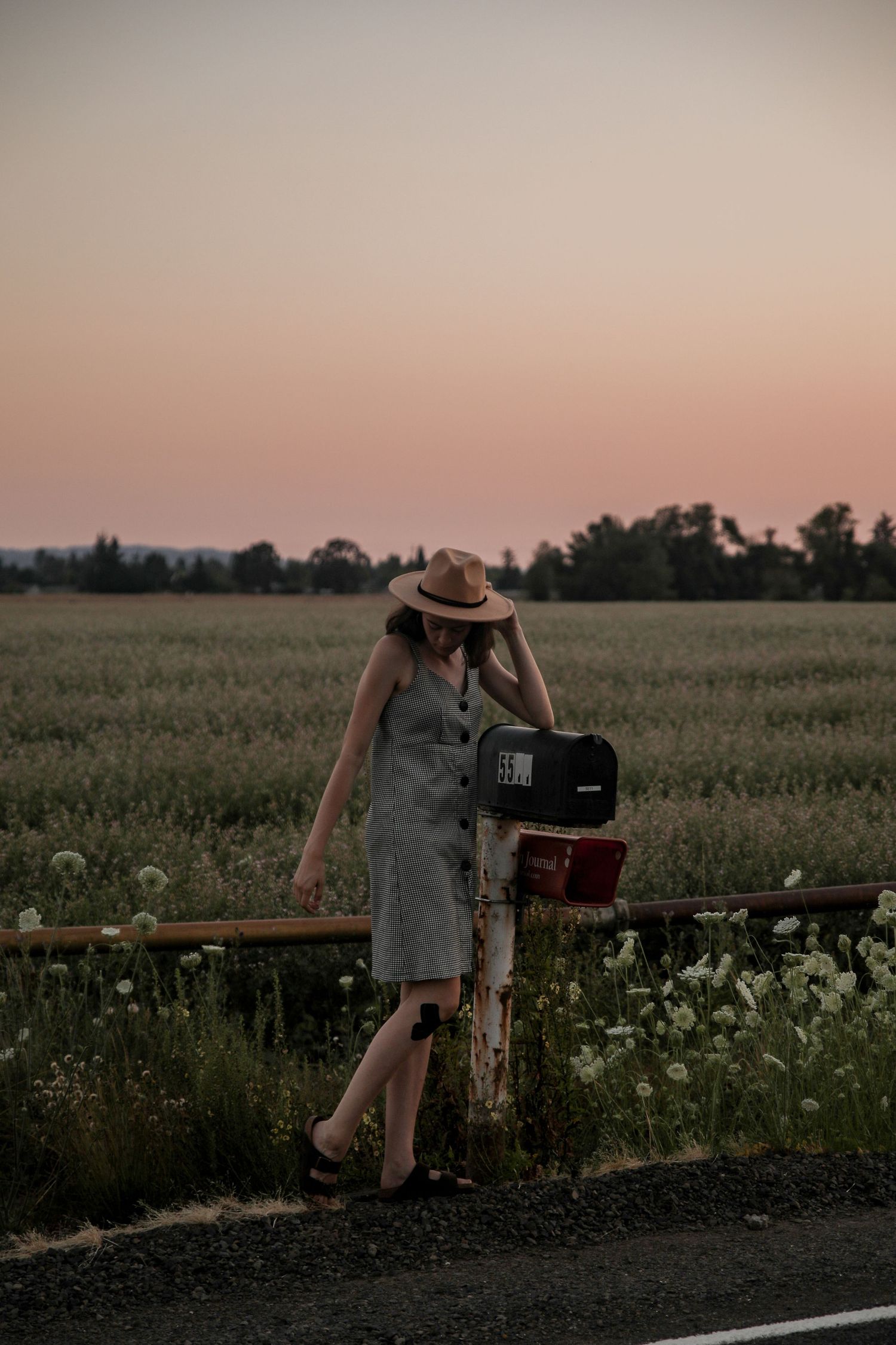 Figure in a checkered dress leans against a post with camera equipment at sunset.