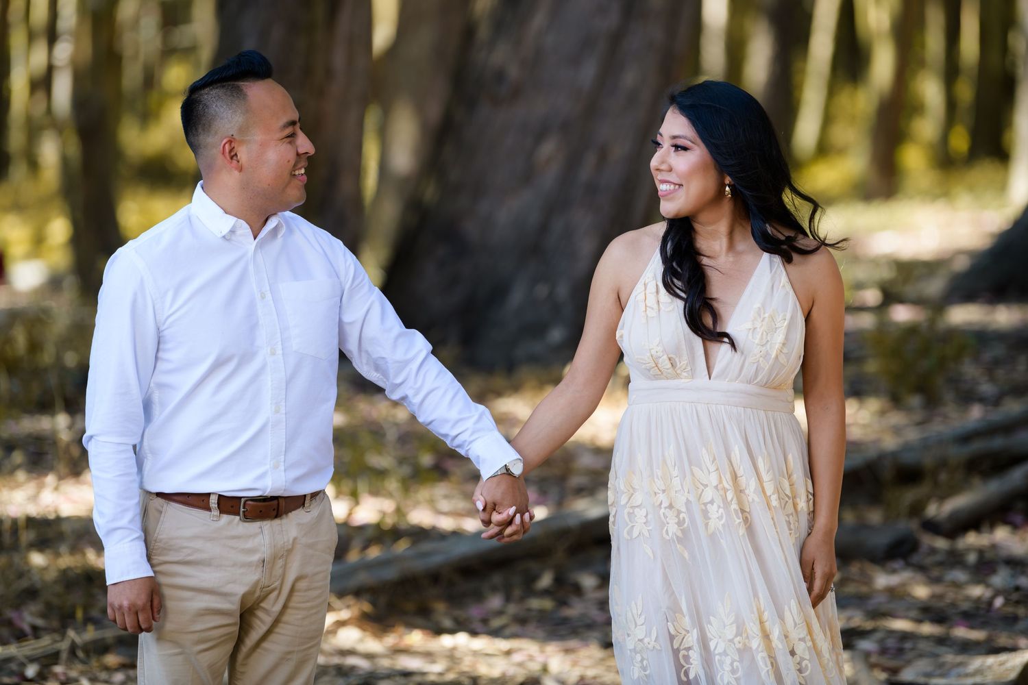 A couple holds hands while walking through a sun-dappled forest, with the woman wearing a flowing cream dress.