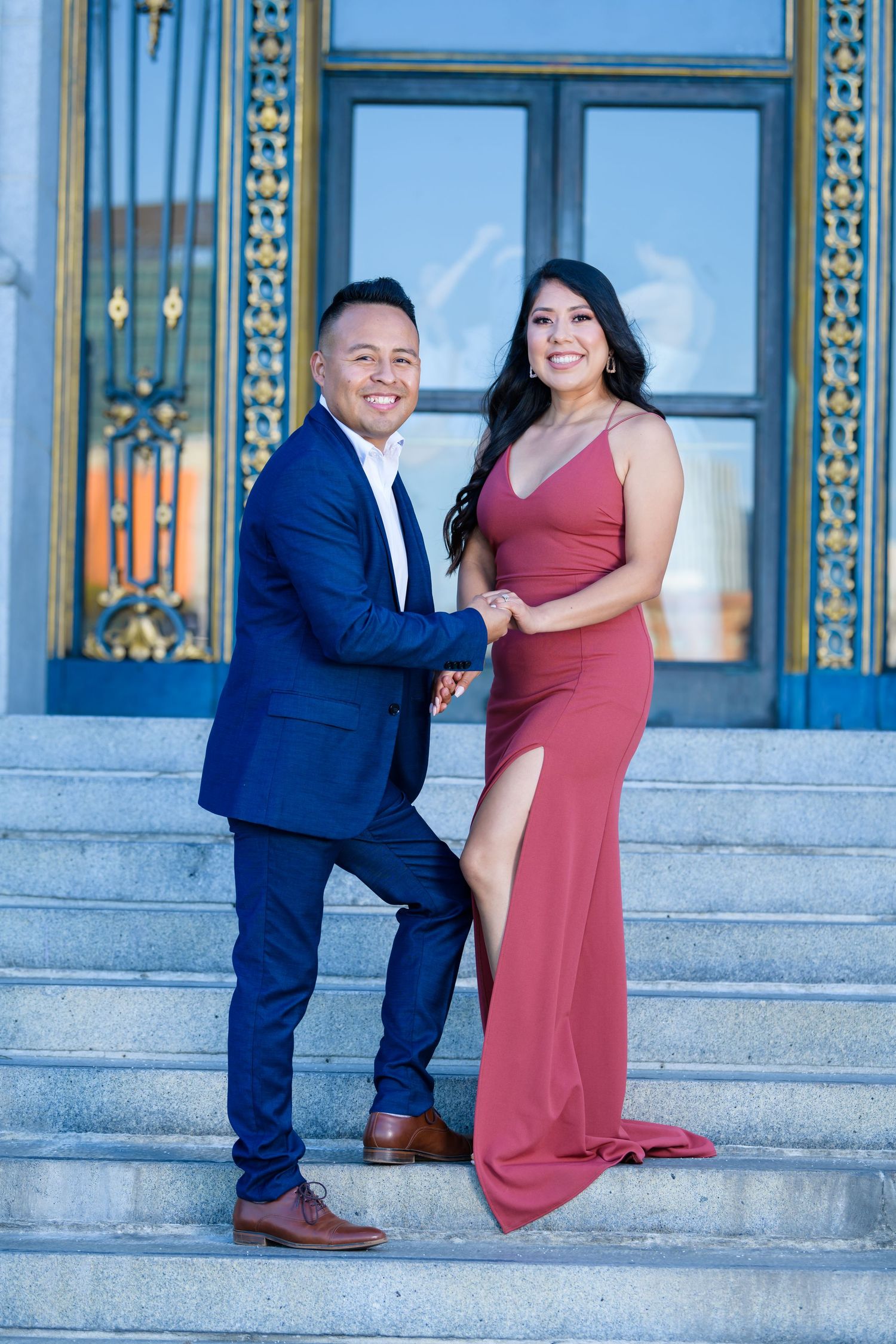 A couple poses on elegant stone steps in formal attire, with a navy suit and burgundy evening gown.