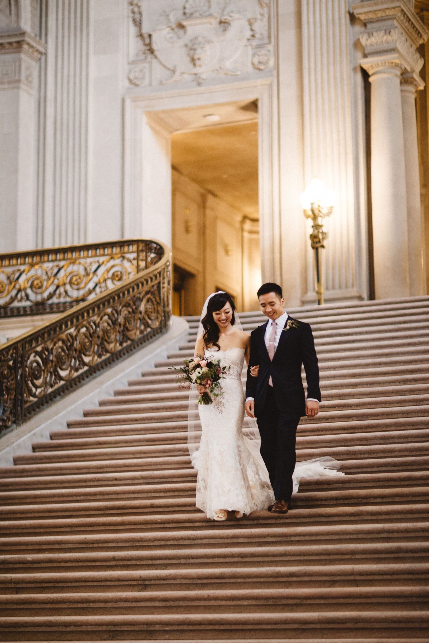 A couple descends an elegant marble staircase with ornate railings in a grand historic building.