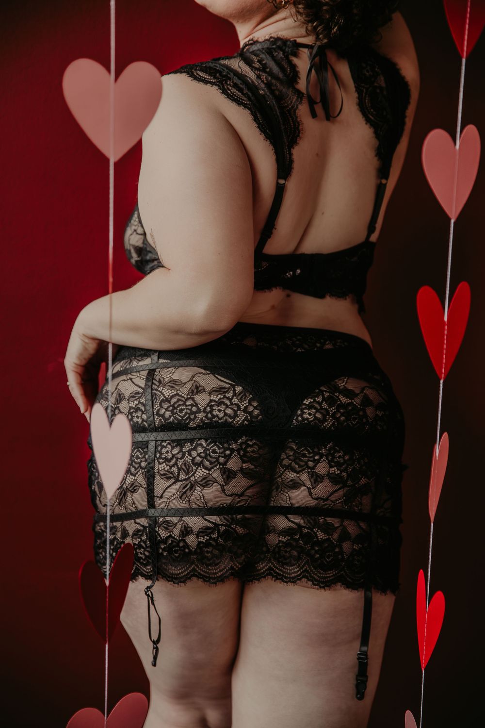Back view of person wearing black lace lingerie against backdrop of hanging heart decorations.