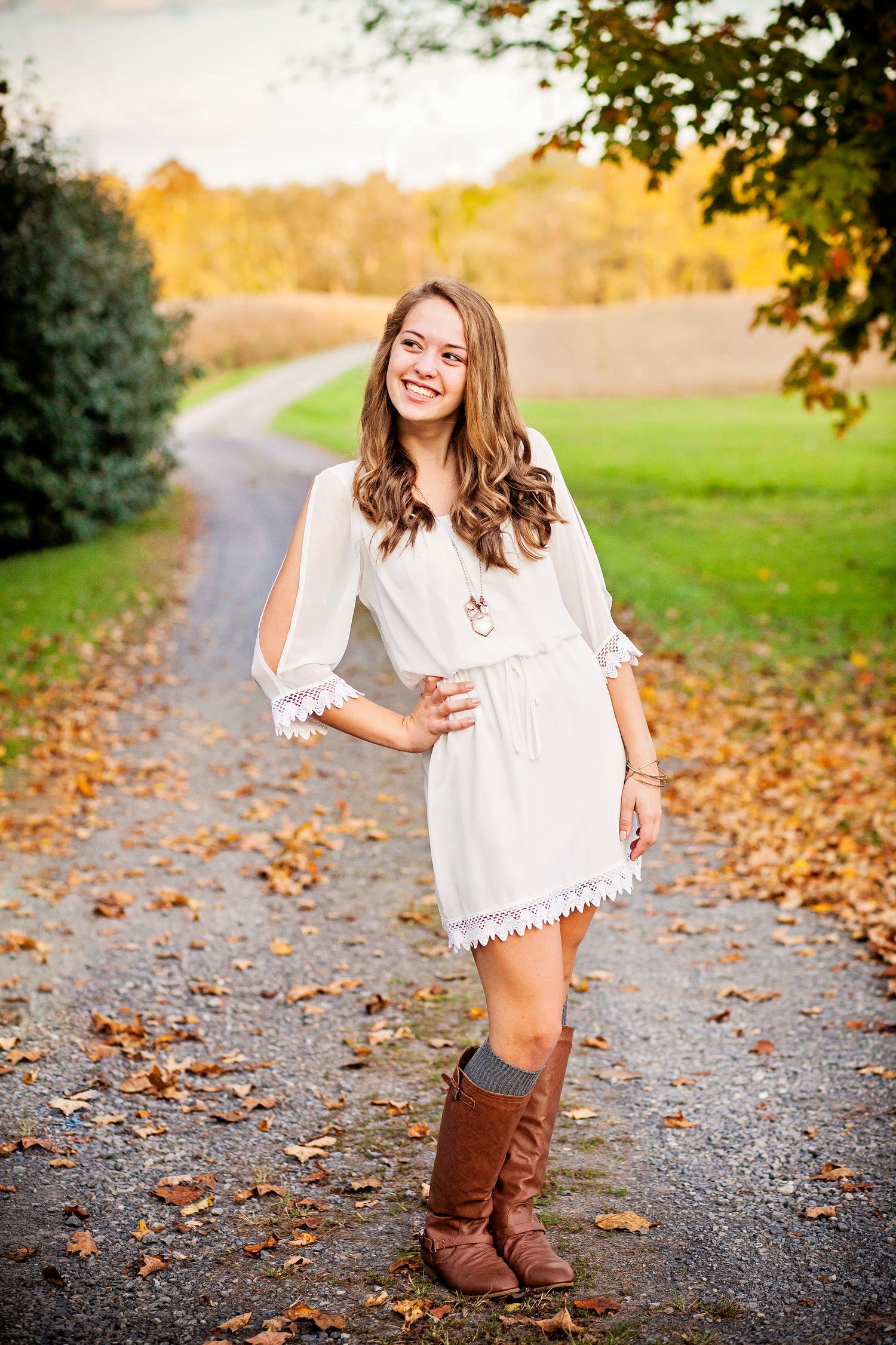 Woman in white dress posing on autumn path with brown boots and falling leaves.