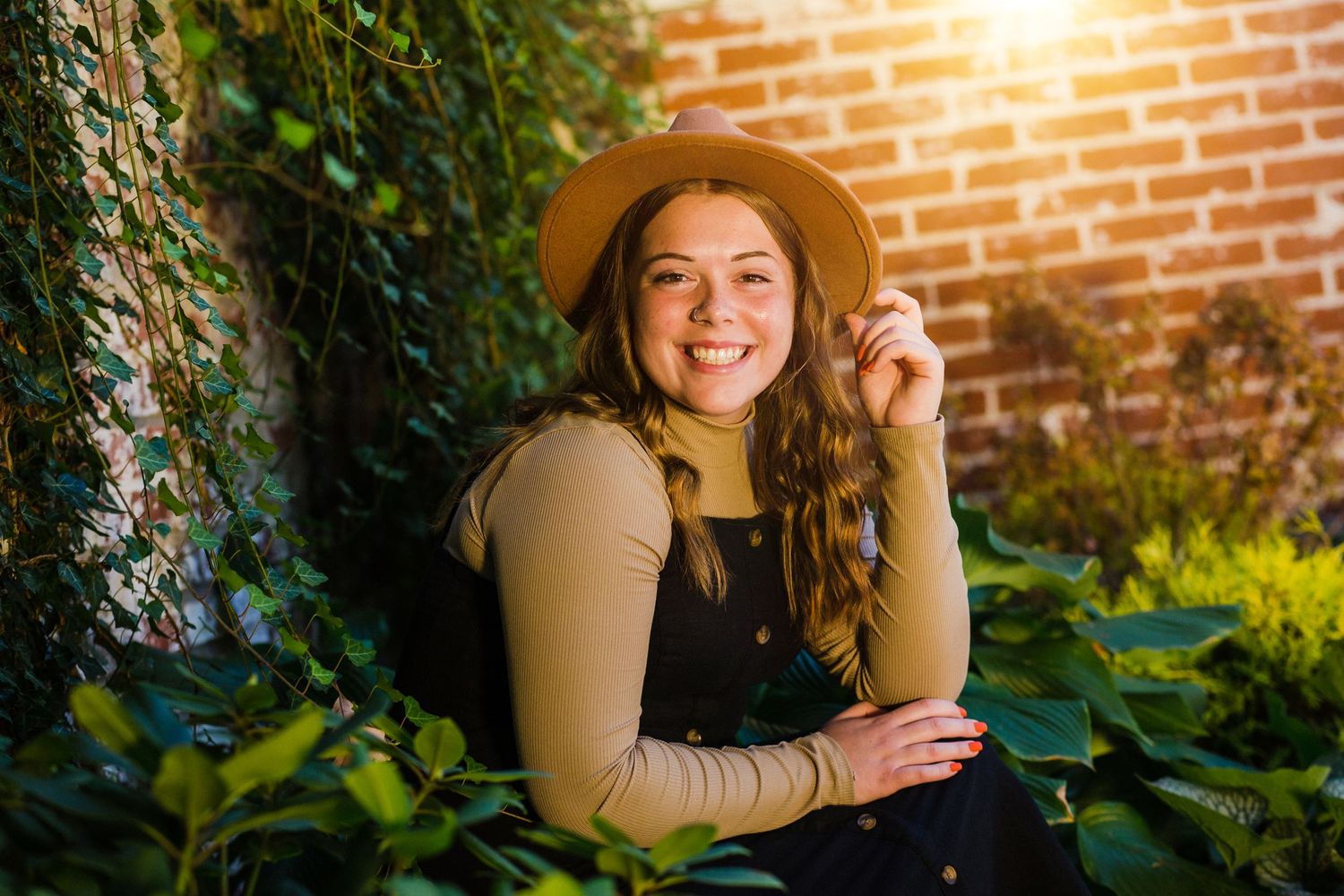A portrait in front of a brick wall with sunlight and greenery wearing a brown hat.
