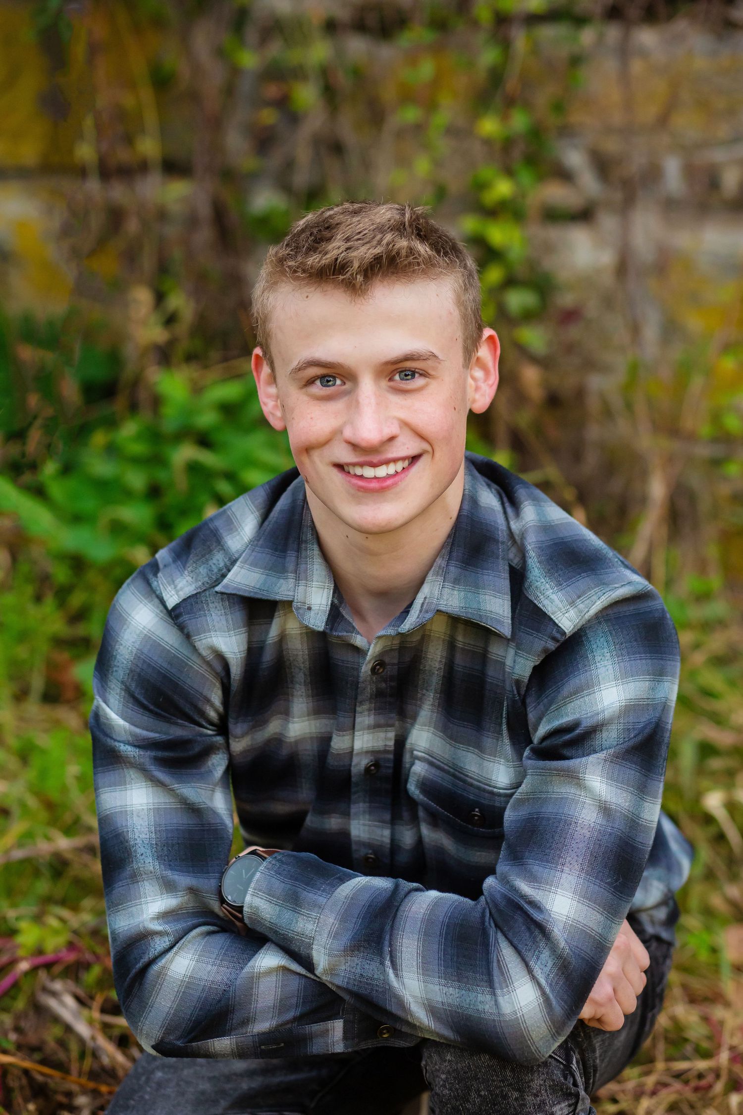 Casual portrait of someone wearing plaid flannel shirt outdoors with natural greenery background.