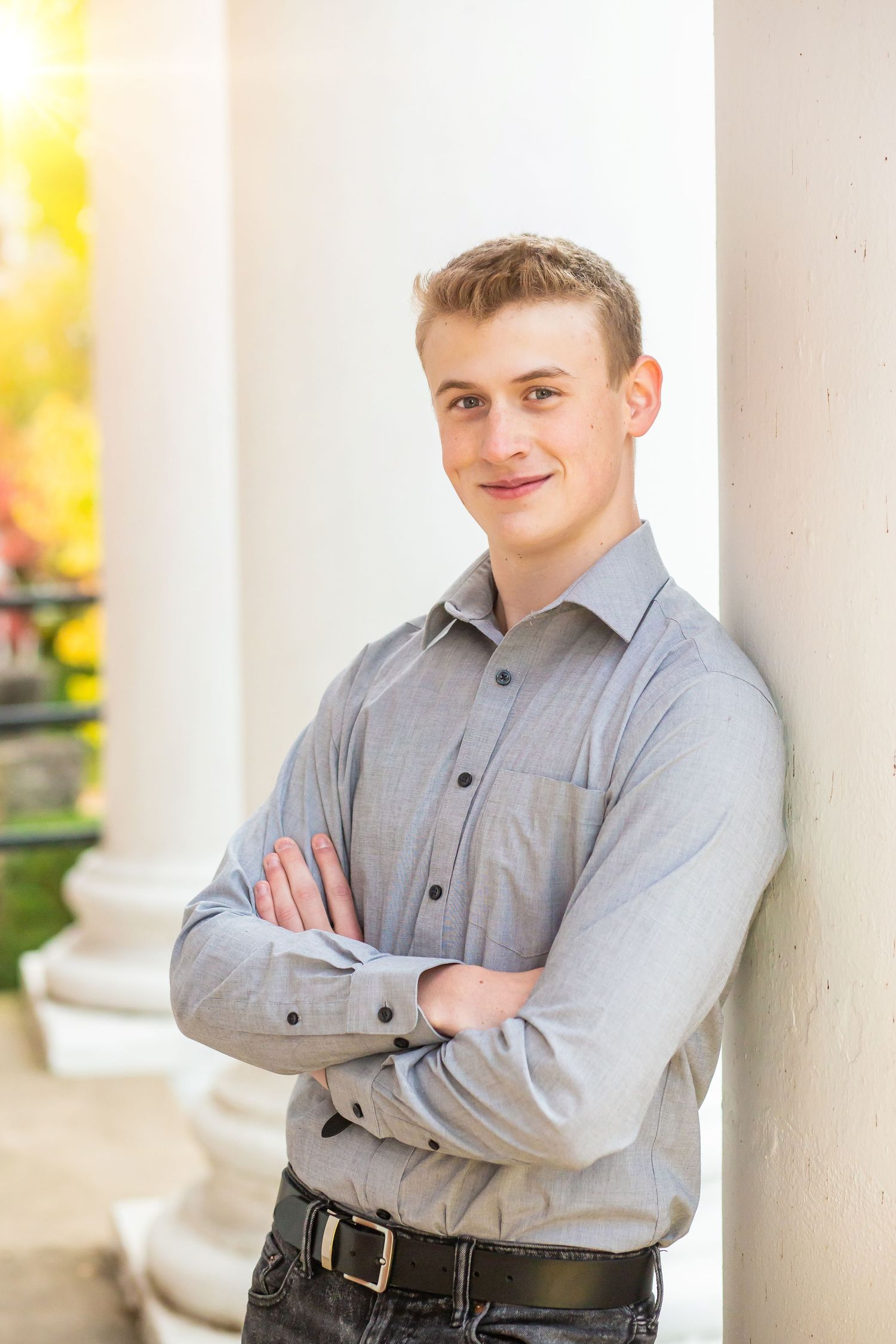 Professional portrait of someone in a grey button-down shirt standing against a white pillar with arms crossed.