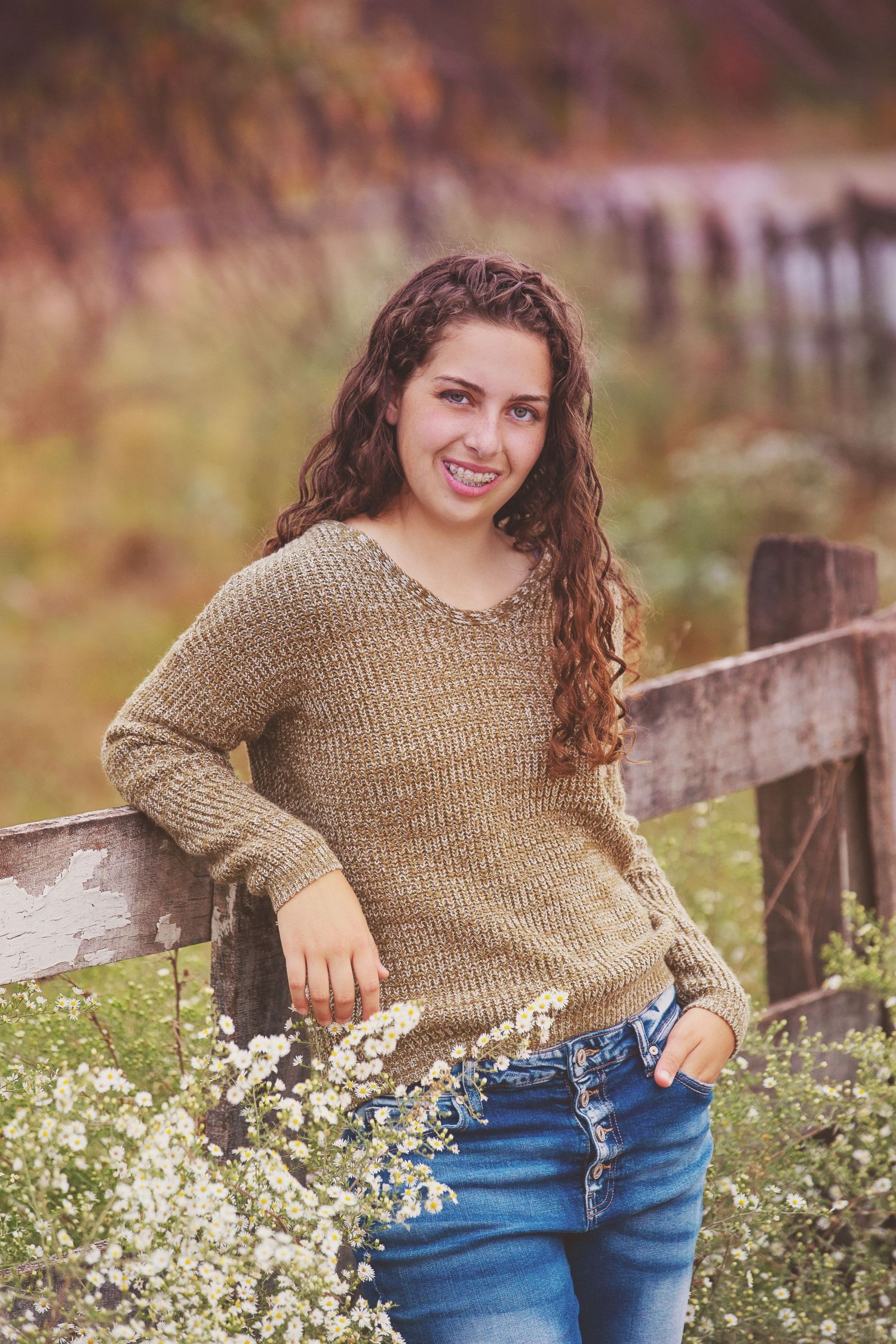 Casual portrait by rustic fence with wildflowers, wearing brown sweater and jeans.