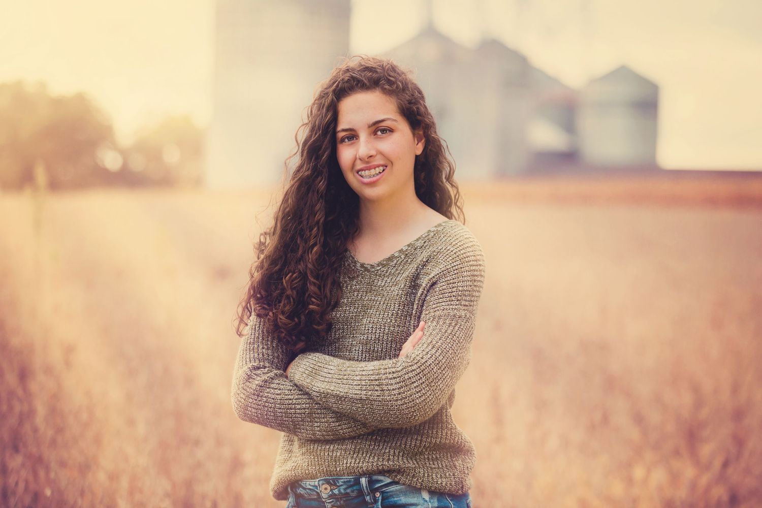 Rural portrait with someone in grey sweater standing in front of grain silos at sunset.