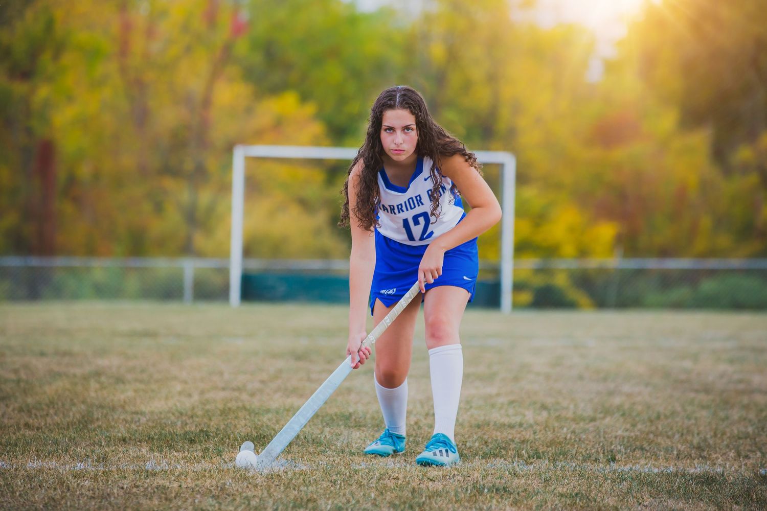 Field hockey player in blue uniform crouching with stick during golden hour on grass field.