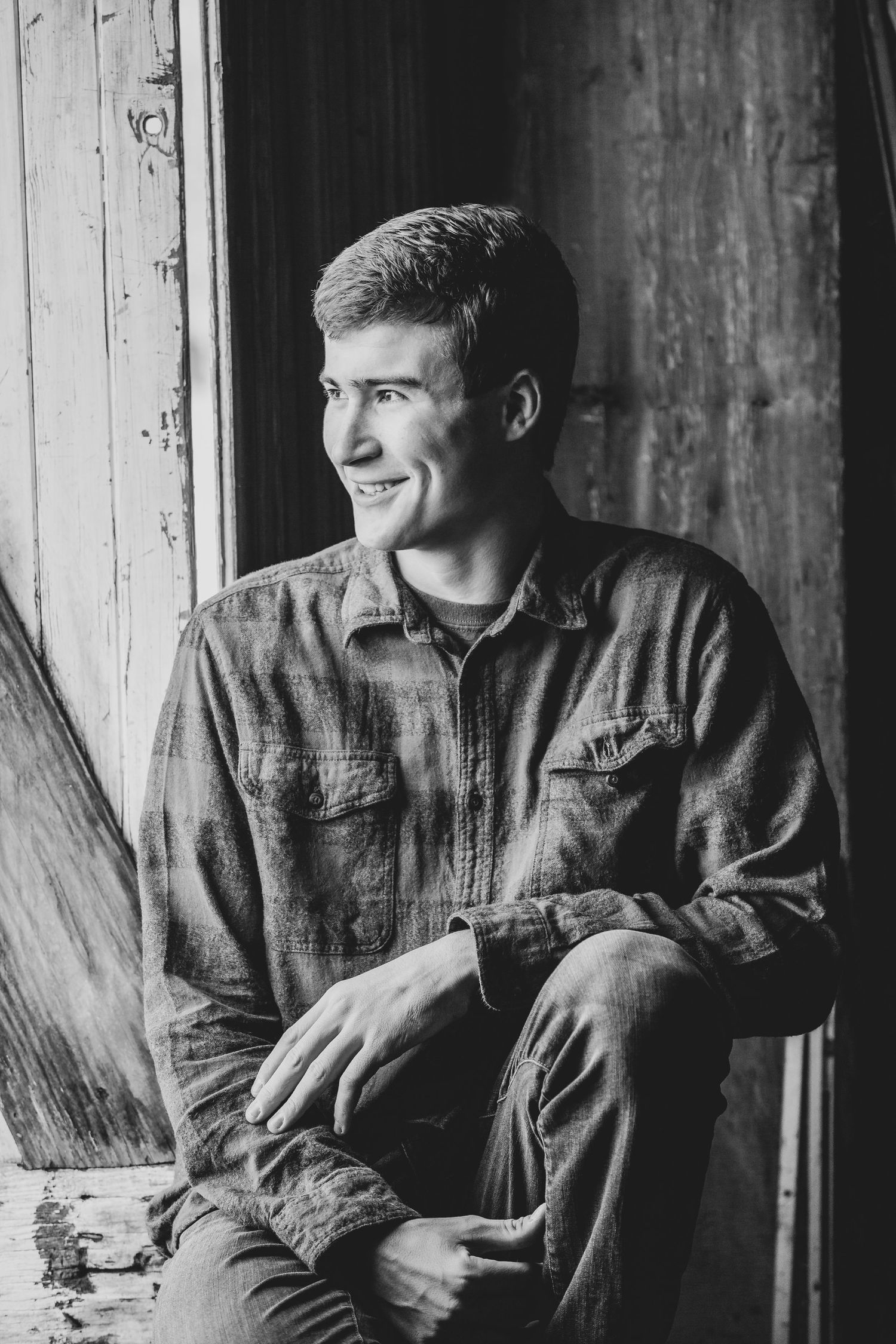 A black and white portrait of someone sitting by a rustic wooden doorway.