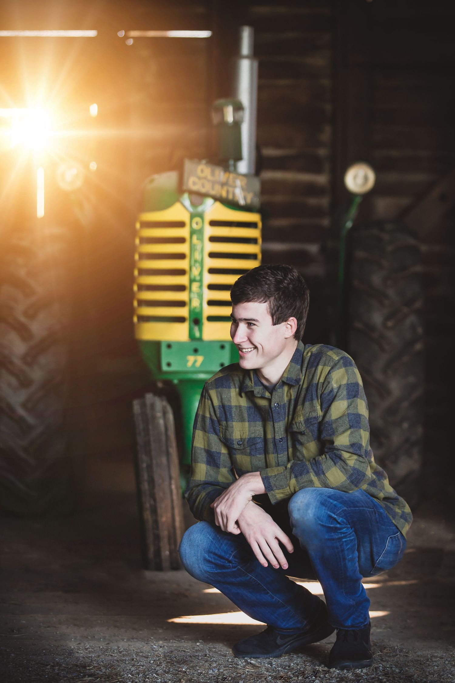 A plaid-shirted figure crouches near a vintage green tractor in a rustic barn setting with dramatic lighting.