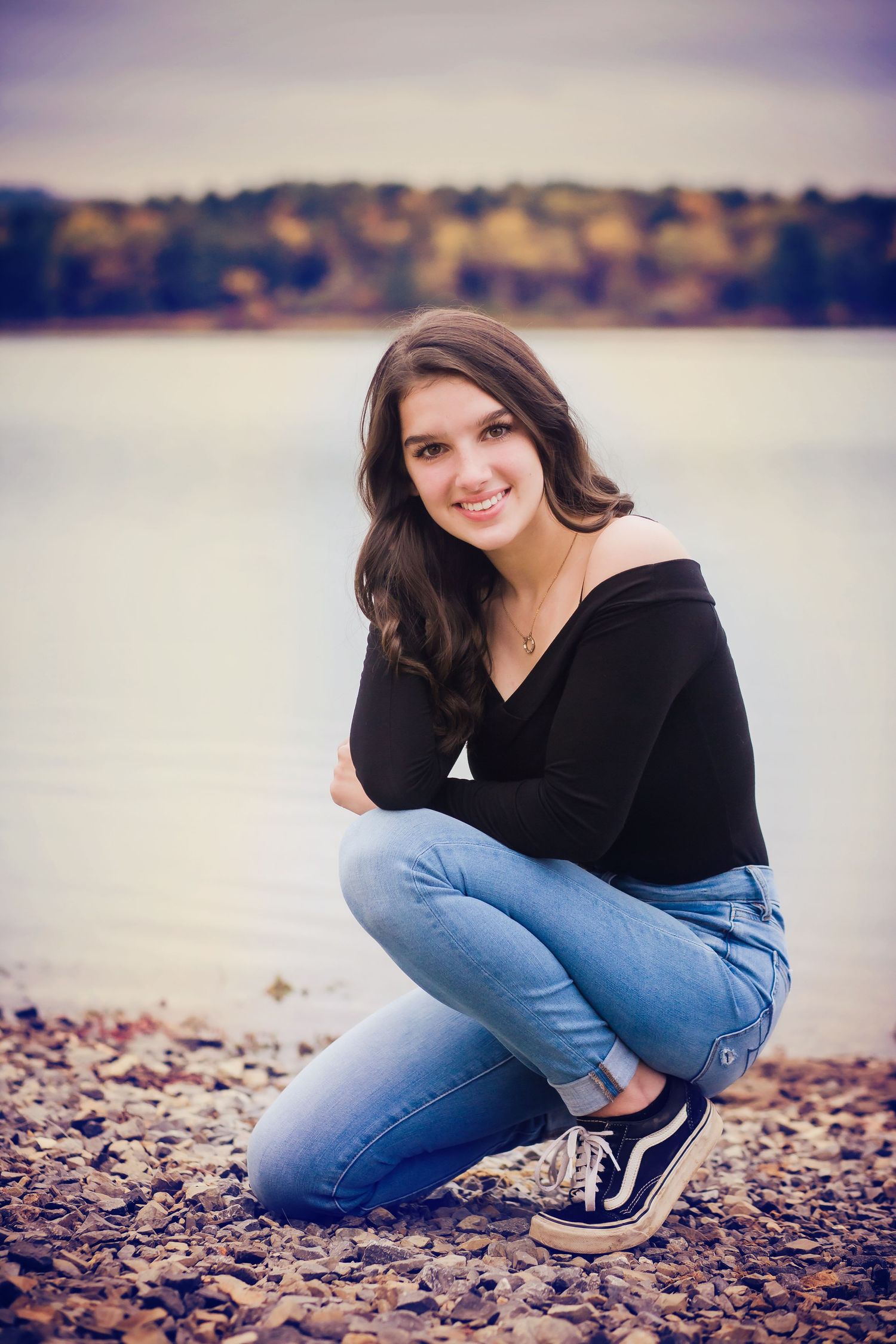 Waterfront portrait with person in black sweater and jeans kneeling on rocky shore.