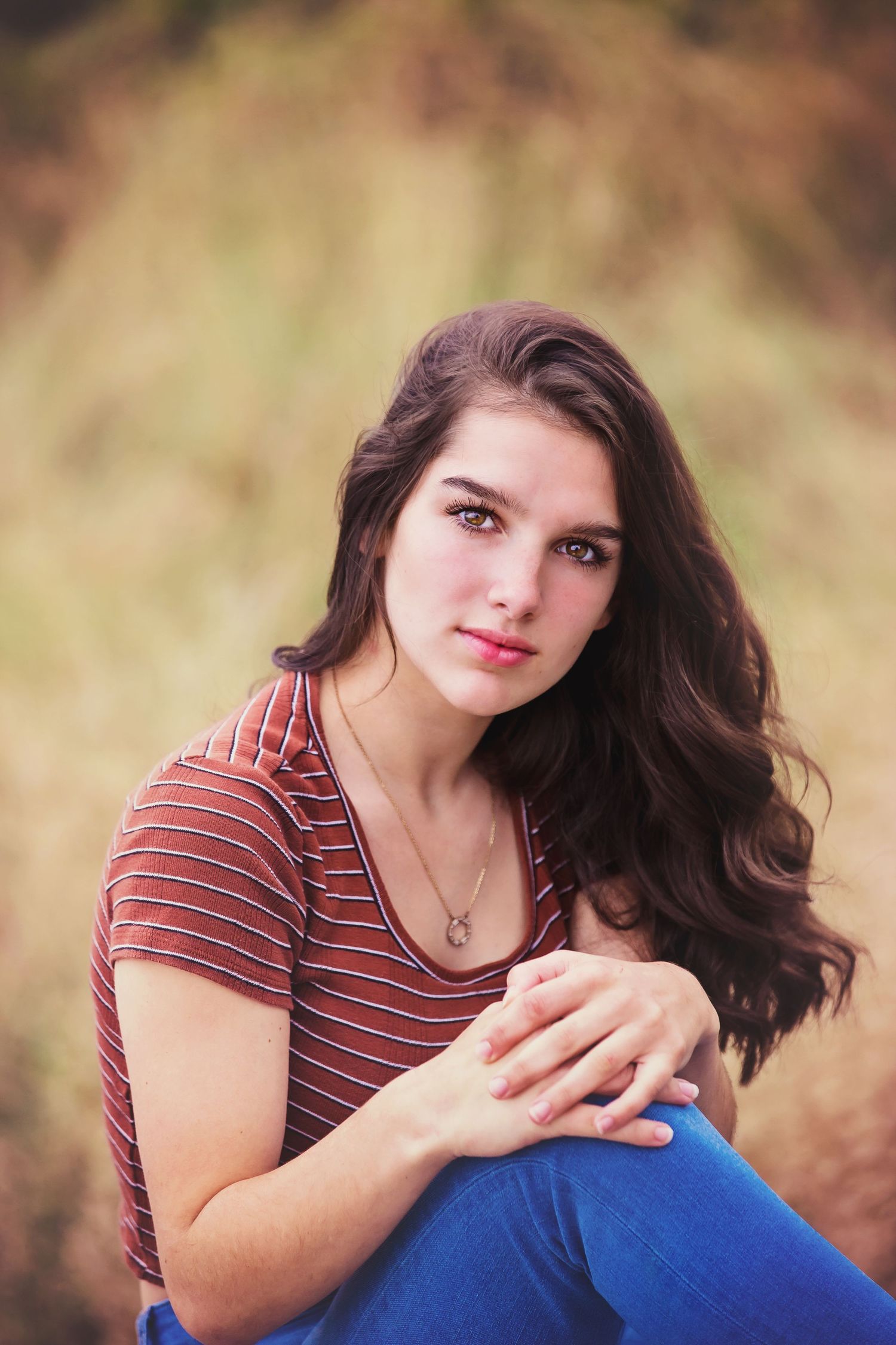 Person in striped shirt and blue jeans poses outdoors with natural autumn backdrop.