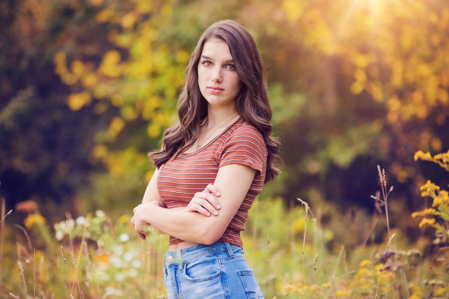 Person in striped shirt and jeans standing in golden sunlit meadow with autumn colors.
