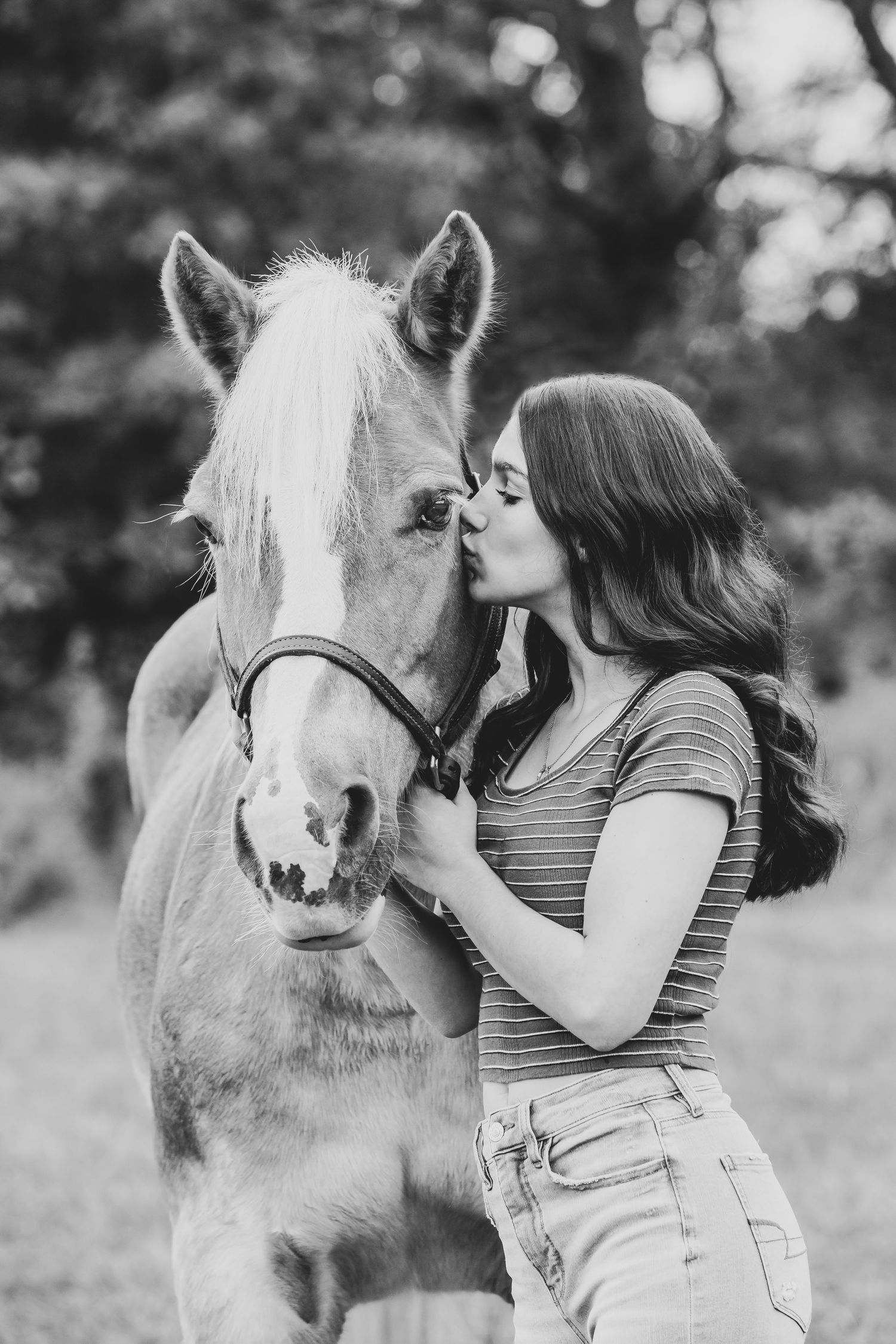 Black and white image of person giving horse a kiss while wearing striped shirt and jeans.