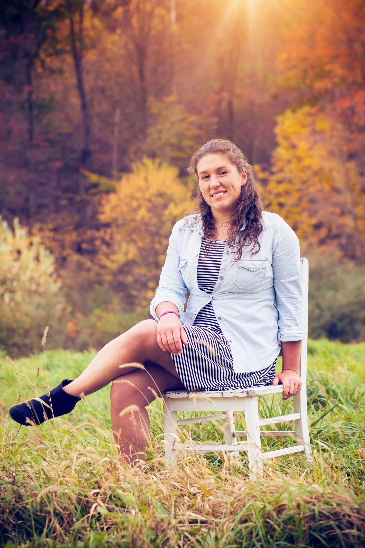 Person in striped dress and light blue shirt poses on white chair in autumn field.