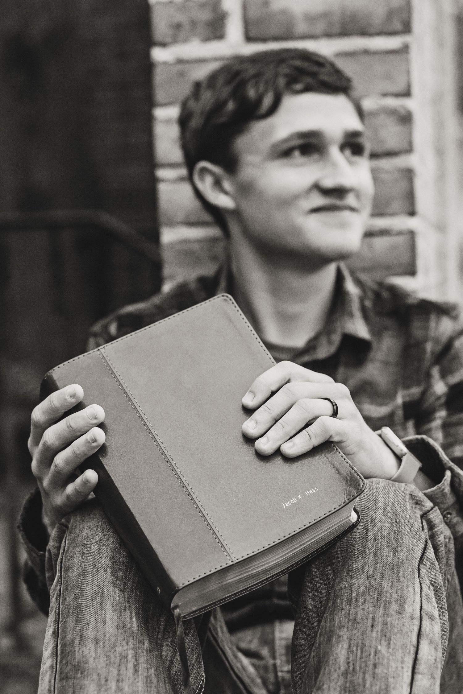 A person holds a leather-bound book while sitting against a brick wall in black and white.