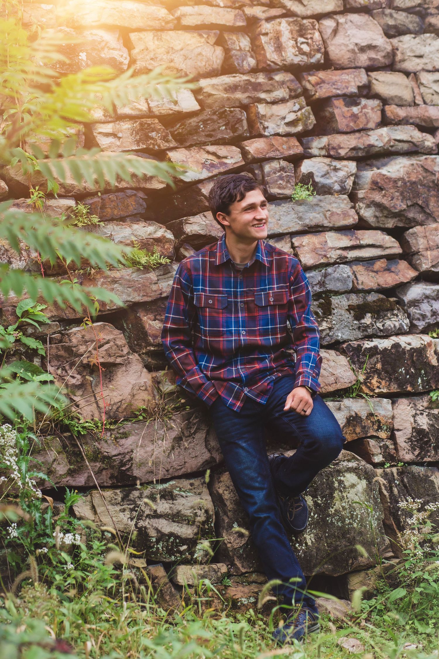 A person in a plaid flannel shirt and jeans leans against a rustic stone wall with sunlight filtering through leaves.