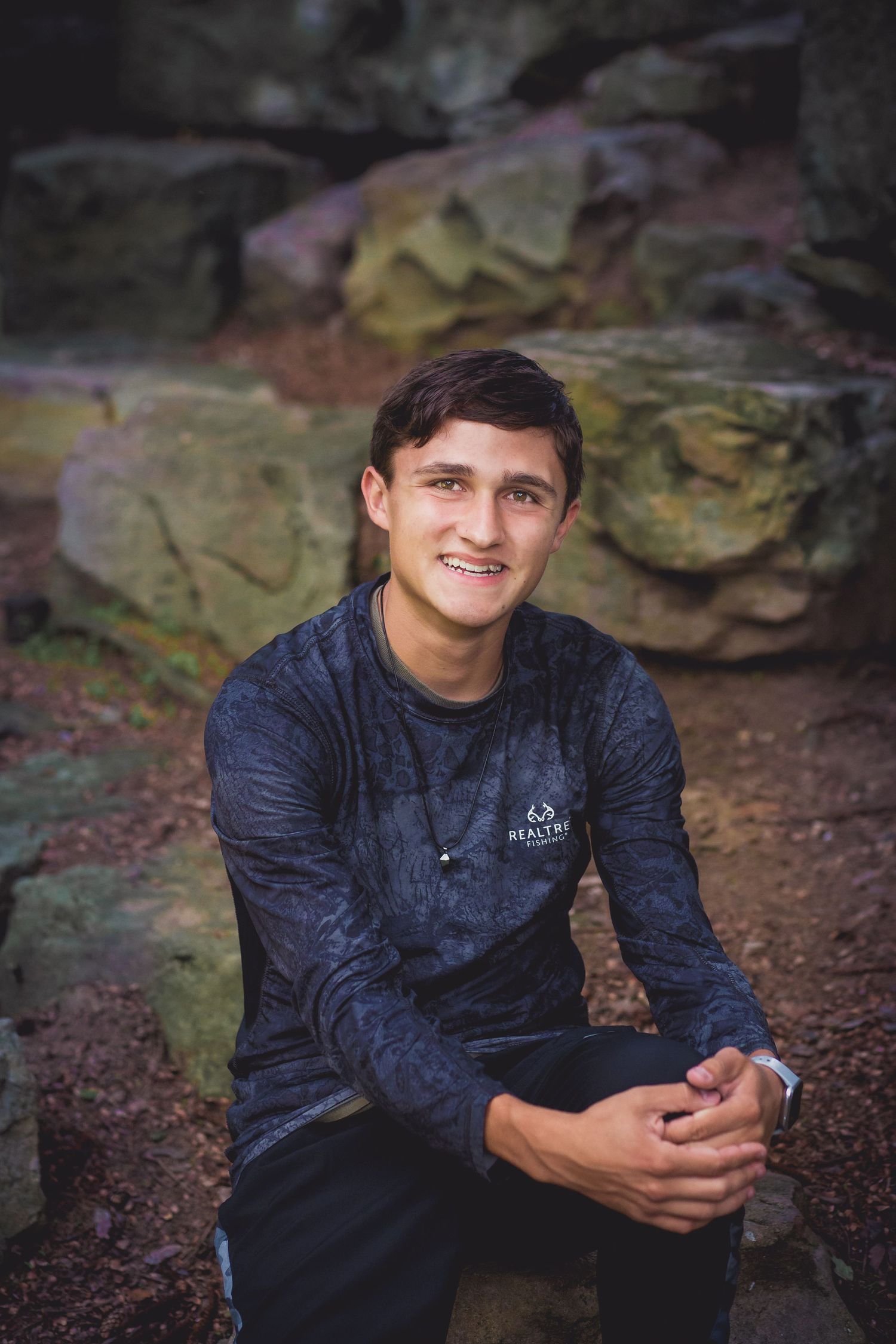 Young person in navy blue sweatshirt sitting on rocky terrain with natural backdrop.